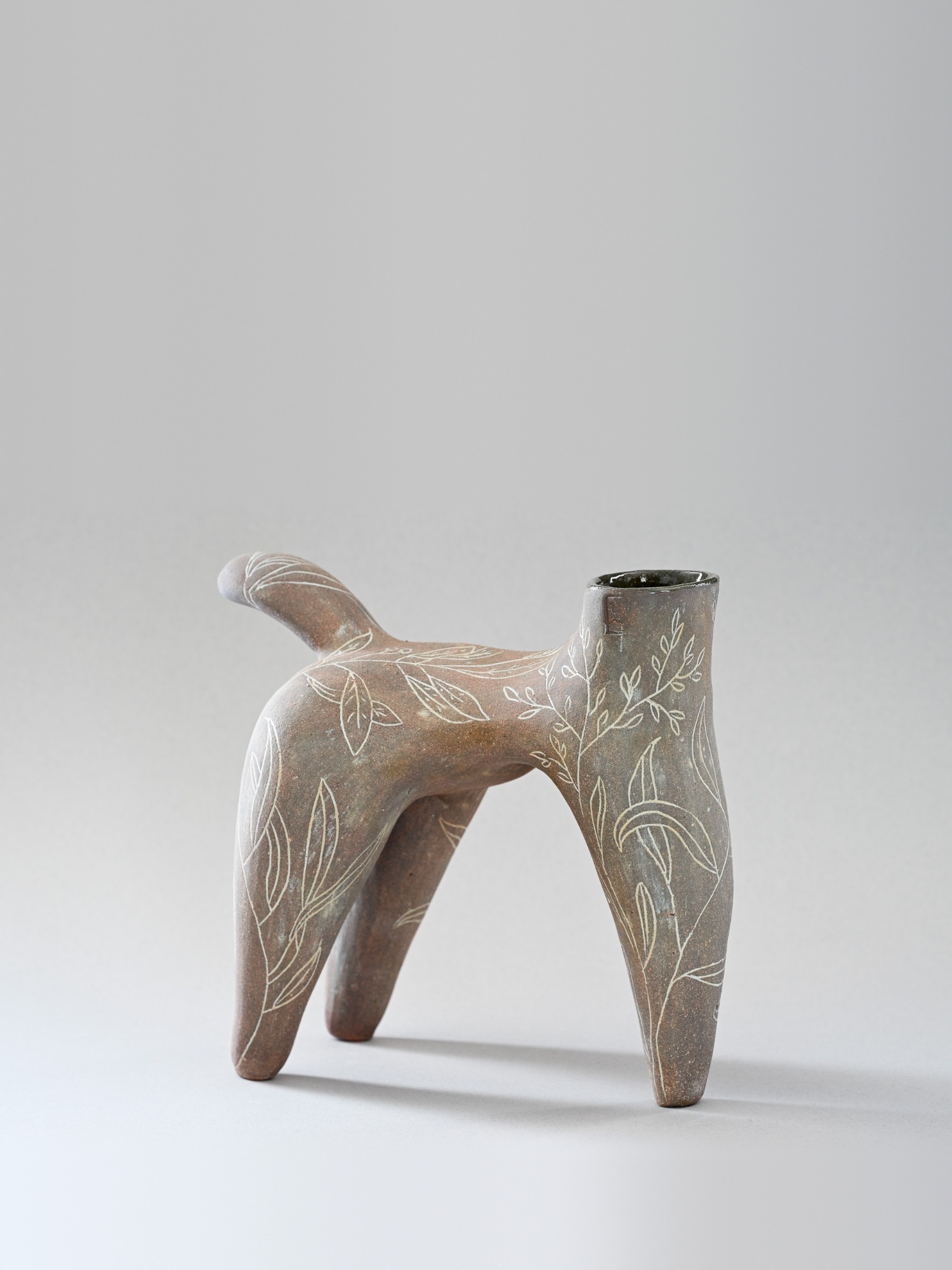 Pièce brune à quatre pieds, au long cou ouvert, couverte de feuillages incisés en blanc, sur fond gris de studio.