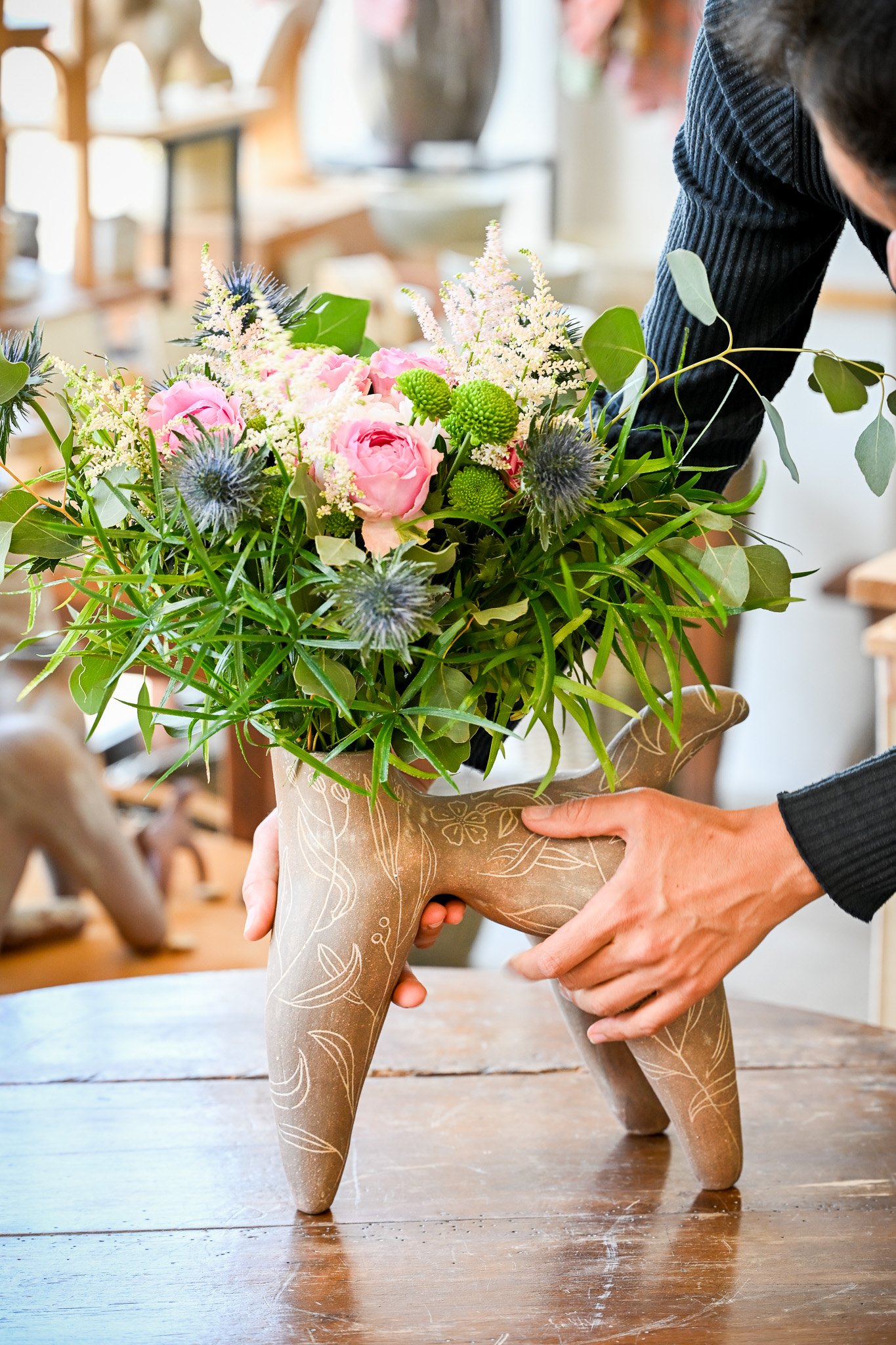 Deux mains posent la pièce sur une table ; un bouquet sort de son col.