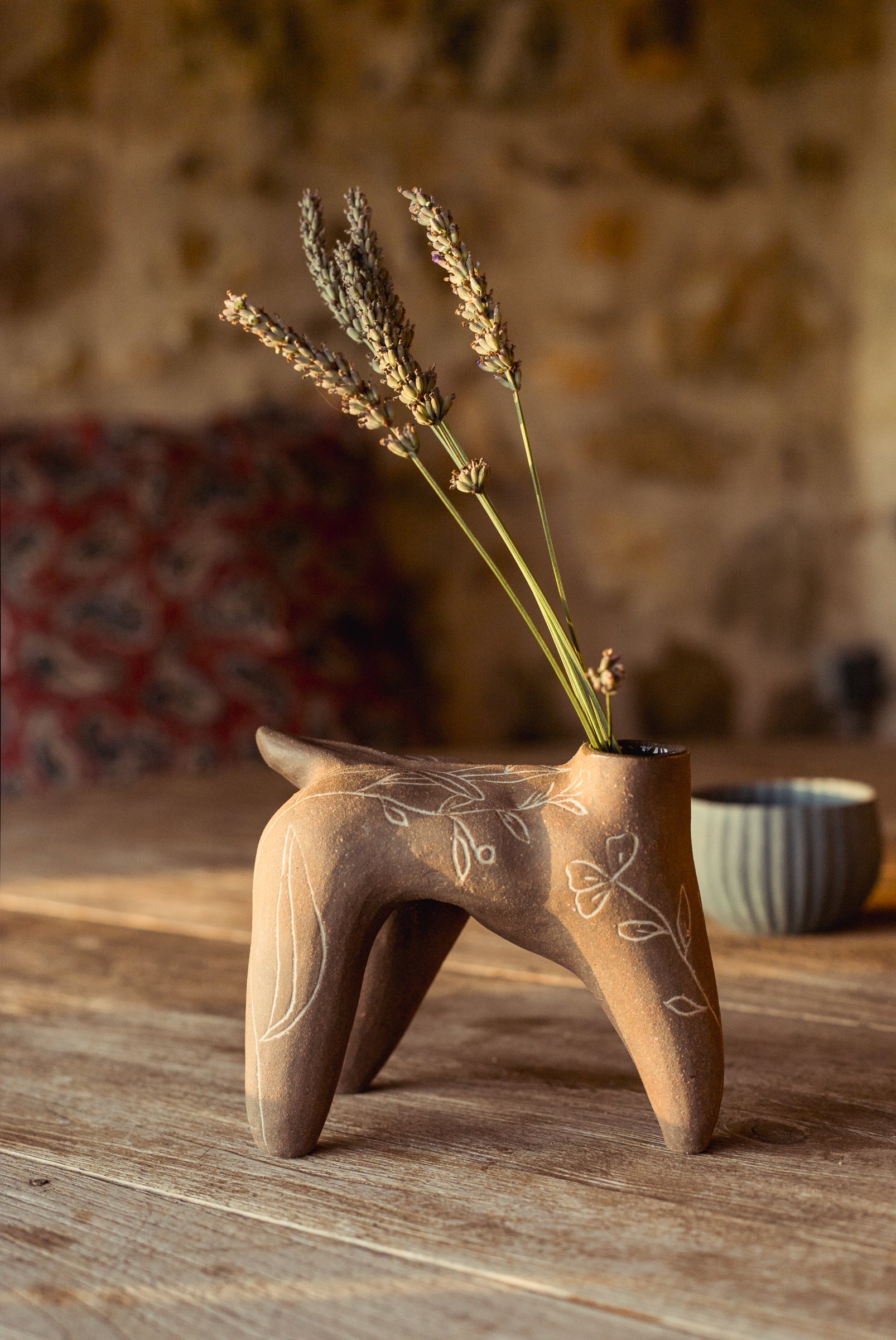 Pièce brune à quatre pieds, vue de biais, des brins de lavande dans son col, sur une table en bois devant un mur de pierre.