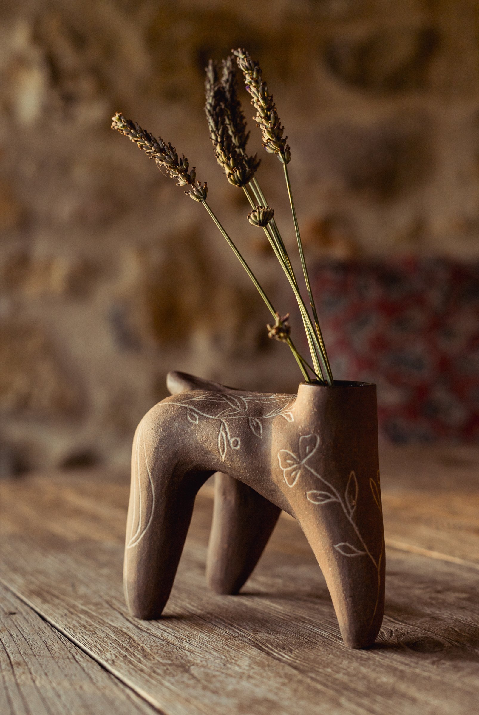 Pièce brune à quatre pieds, feuillages incisés, tenant des épis de graminées séchées, sur une table en bois devant un mur de pierre.