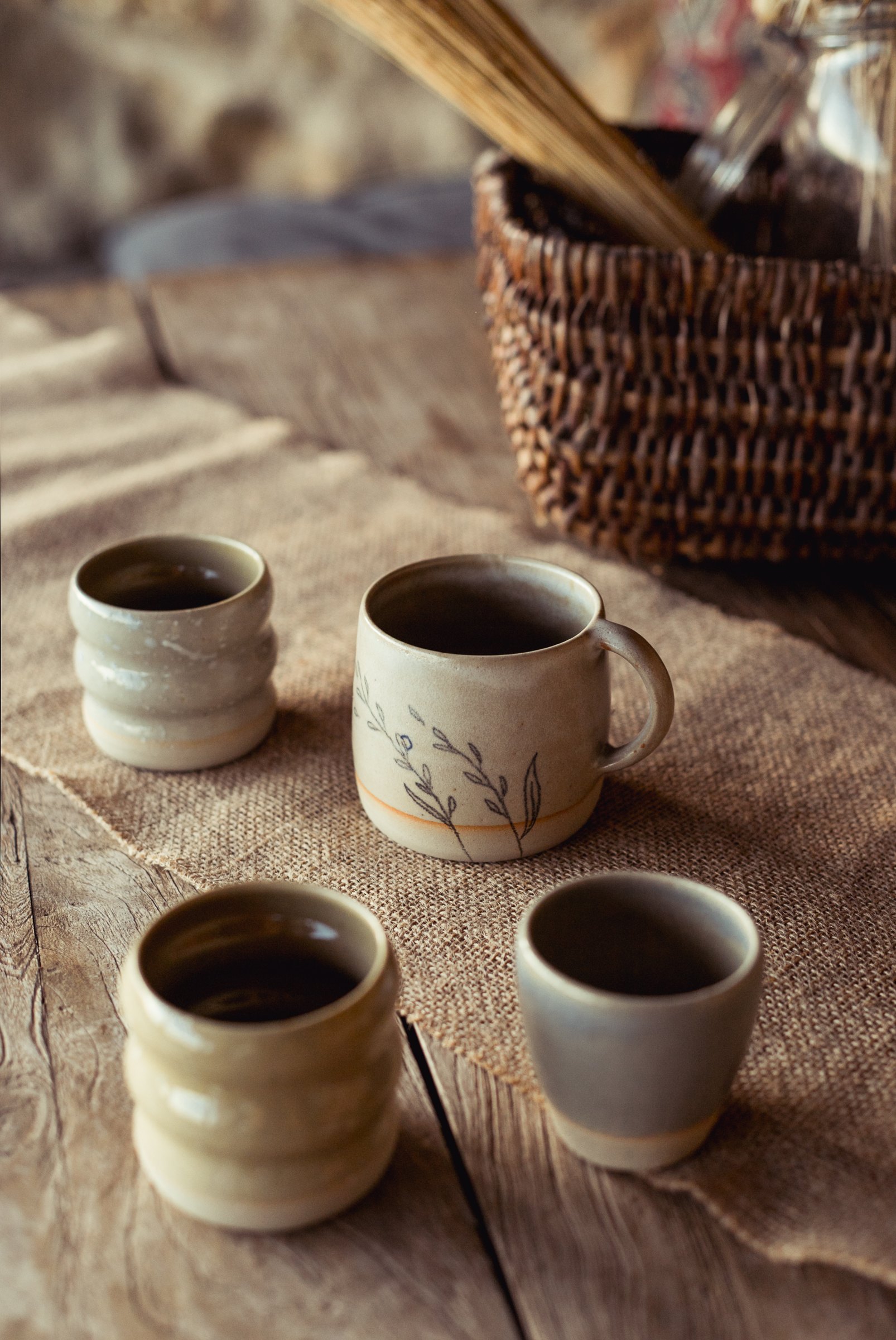 Trois tasses et un gobelet aux émaux clairs, posés sur une toile de lin, panier en osier derrière.