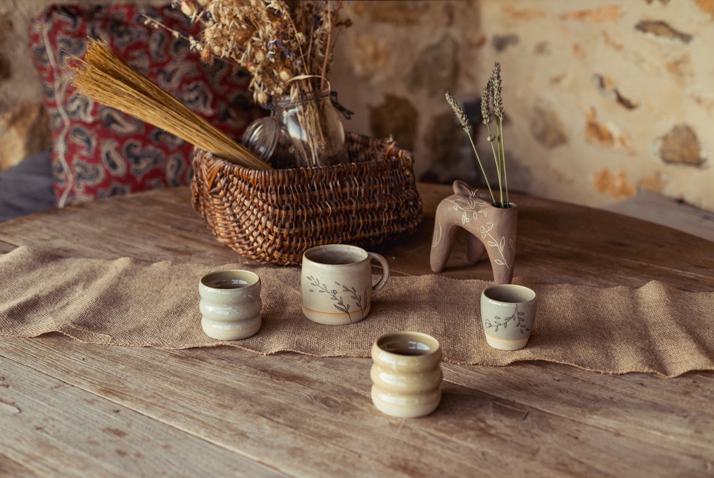 Table en bois : quatre tasses claires et une pièce brune à quatre pieds sur une toile de lin, panier de fleurs séchées, mur de pierre.