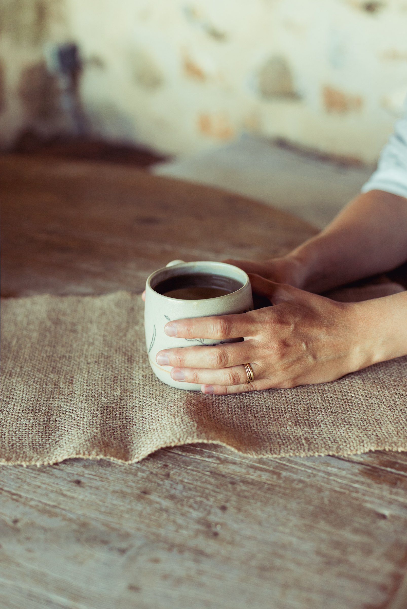 Deux mains entourent une tasse claire décorée d'un brin, sur une toile de lin.