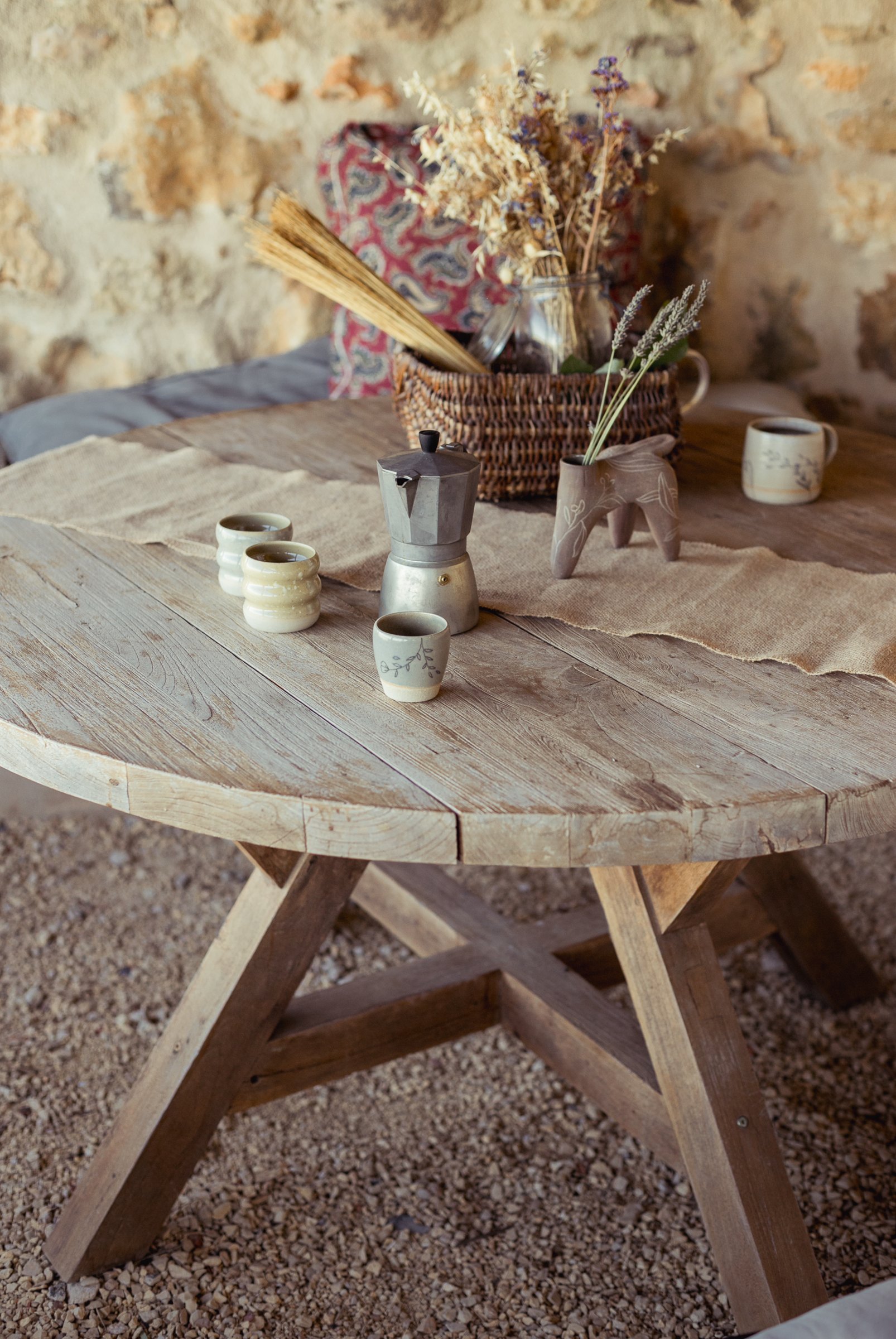 Table ronde en bois avec des tasses et un panier de fleurs séchées, devant un mur de pierre.