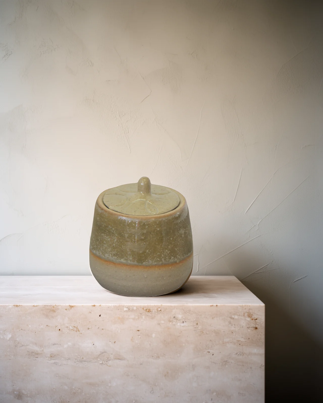 Petit pot à couvercle en grès, émail olive et crème, sur un socle de travertin pâle.
