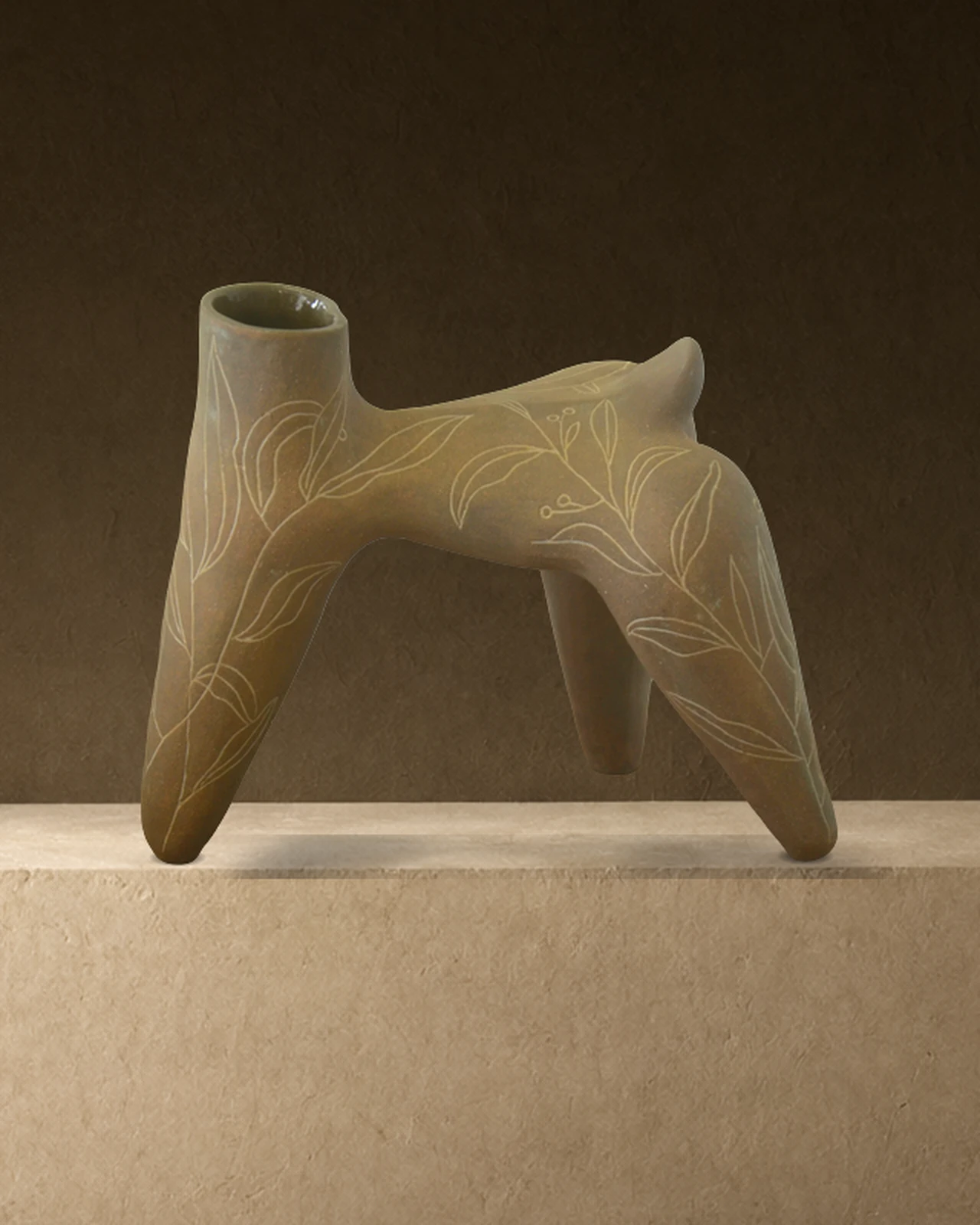 Vase à quatre pattes en terre brune, gravé de feuillages, posé sur un socle bas devant un mur sombre.