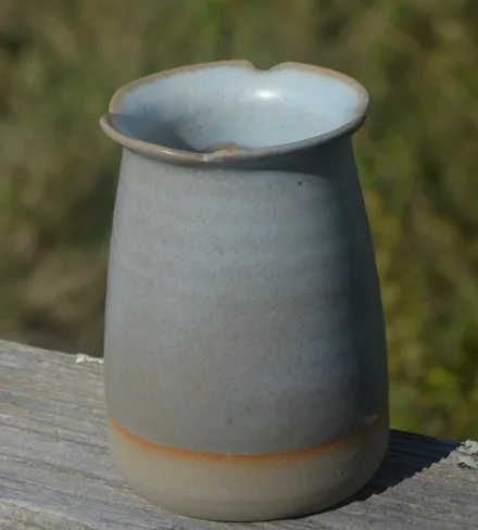 Vase en grès bleu-gris au bord festonné, posé sur une barrière en bois devant un pré.