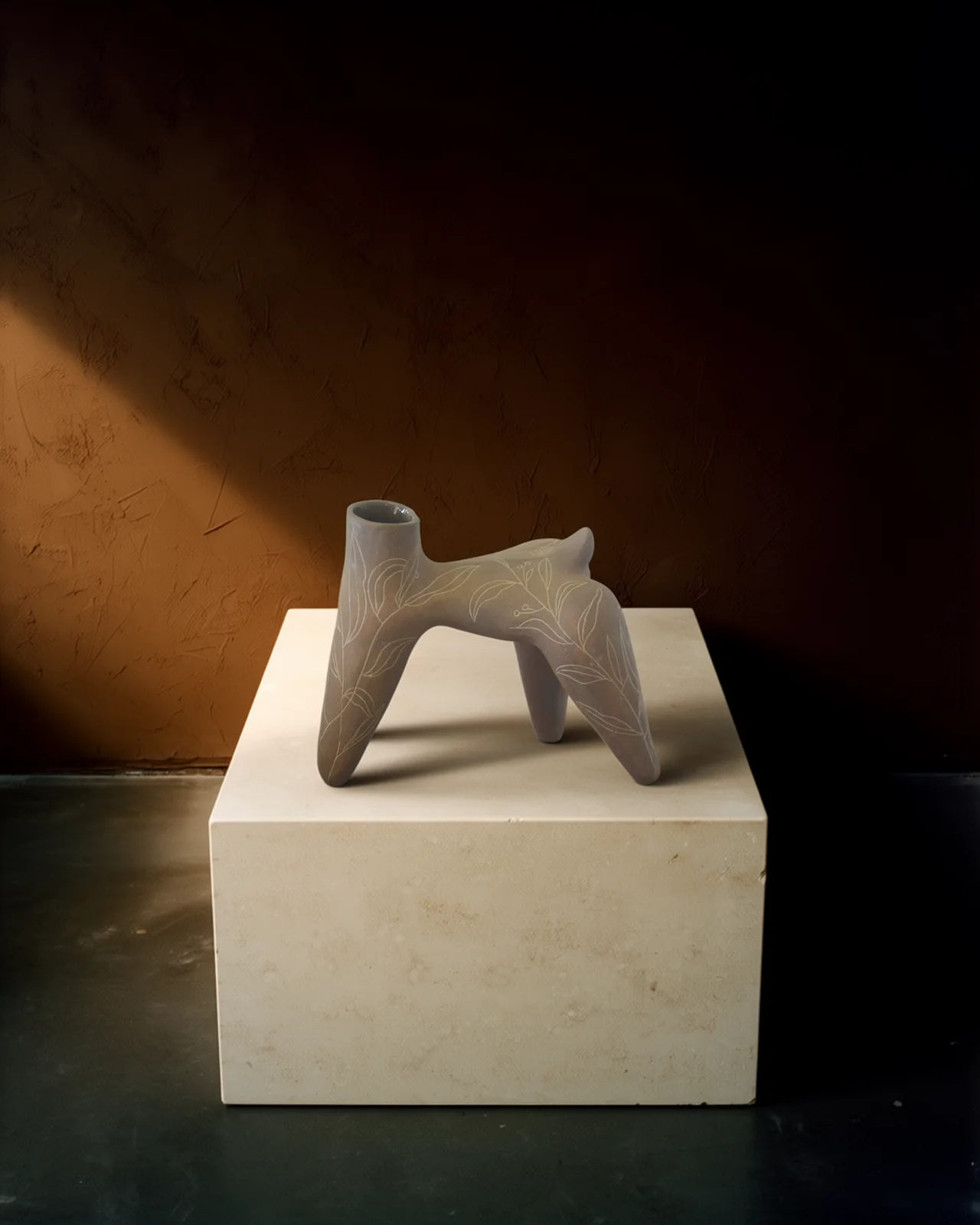Vase à quatre pattes en terre brune, gravé de feuillages, posé sur un socle de calcaire clair, éclairé de la gauche