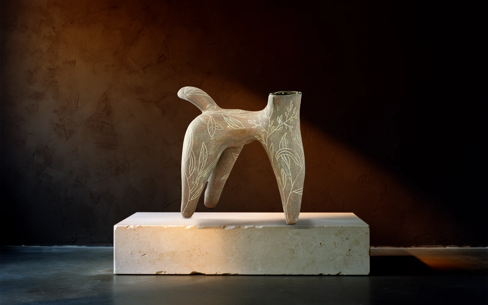 Sculpture à quatre pattes gravée de feuillages, posée sur un socle de travertin, éclairée de la gauche